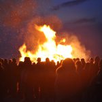 Eine große Menschenmenge steht im Dunkeln um ein helles, großes Feuer, das Funken in die Luft sprüht. Der Nachthimmel ist im Hintergrund leicht bewölkt.