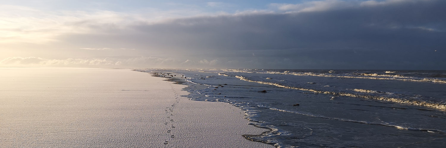 Biikebrennen In Sankt Peter Ordingnkt Peter Ording 3 Weitläufiger Strand bei Sonnenaufgang. Die Oberfläche wirkt verschneit, das Meer schimmert grau-blau. Am Himmel ziehen dichte Wolken auf, während die Sonne links am Horizont einen warmen Schein erzeugt. Fußspuren verlaufen parallel zum Wasser.
