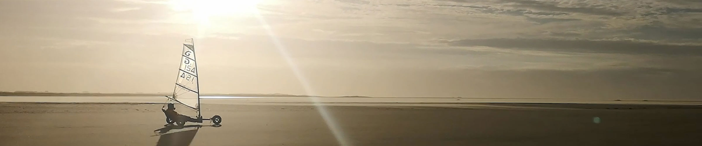 Twilling Spo Schnapper Angebot Header Strandsegler Eine Person fährt mit einem Strandsegler über einen weiten Sandstrand. Die Sonne steht tief am Himmel und wirft lange Schatten. Der Himmel ist leicht bewölkt, und das Wasser ist in der Ferne zu sehen.