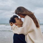 Ein Paar am Strand. Die Frau umarmt den Mann und blickt ihm lächelnd in die Augen. Im Hintergrund sind das Meer und ein bewölkter Himmel zu sehen.